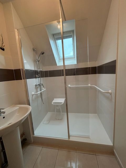 Douche avec siège, rampes et plafond en pente dans une salle de bains avec puits de lumière et lavabo.