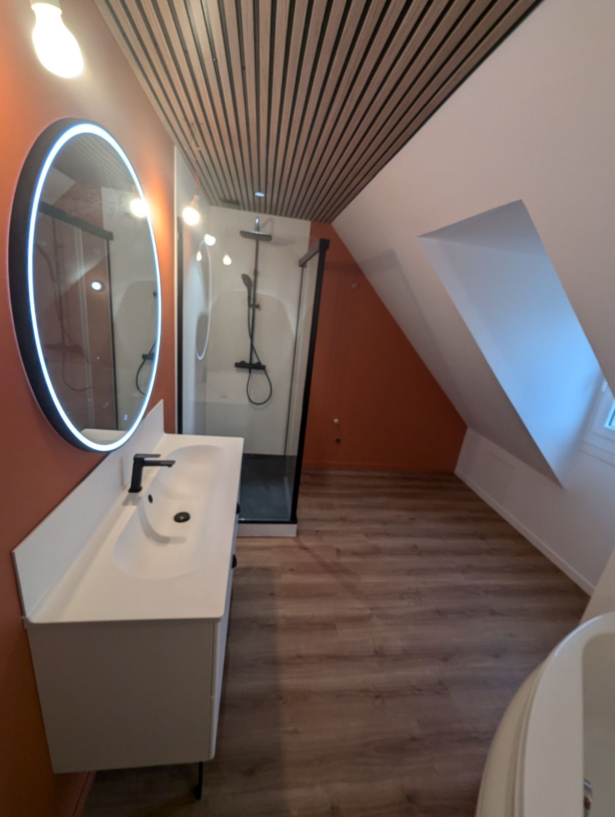 Salle de bains avec vanité blanche, douche noire, murs orange, plafond en pente avec panneaux de bois et un grand miroir ovale.