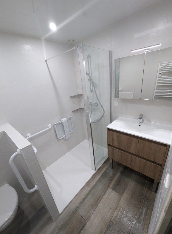 Salle de bains accessible avec murs blancs, douche en verre, vanité en bois et barres d'appui.