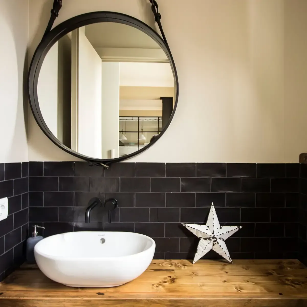 Baño con espejo redondo, azulejo negro, lavabo blanco y encimera de madera.
