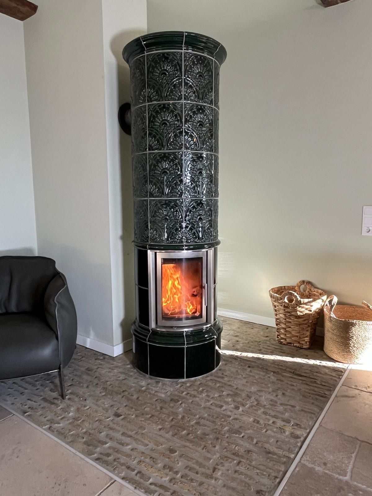 Ein hoher, grün gekachelter Holzofen, aus dem Flammen sichtbar sind. Daneben ein schwarzer Stuhl und Weidenkörbe.