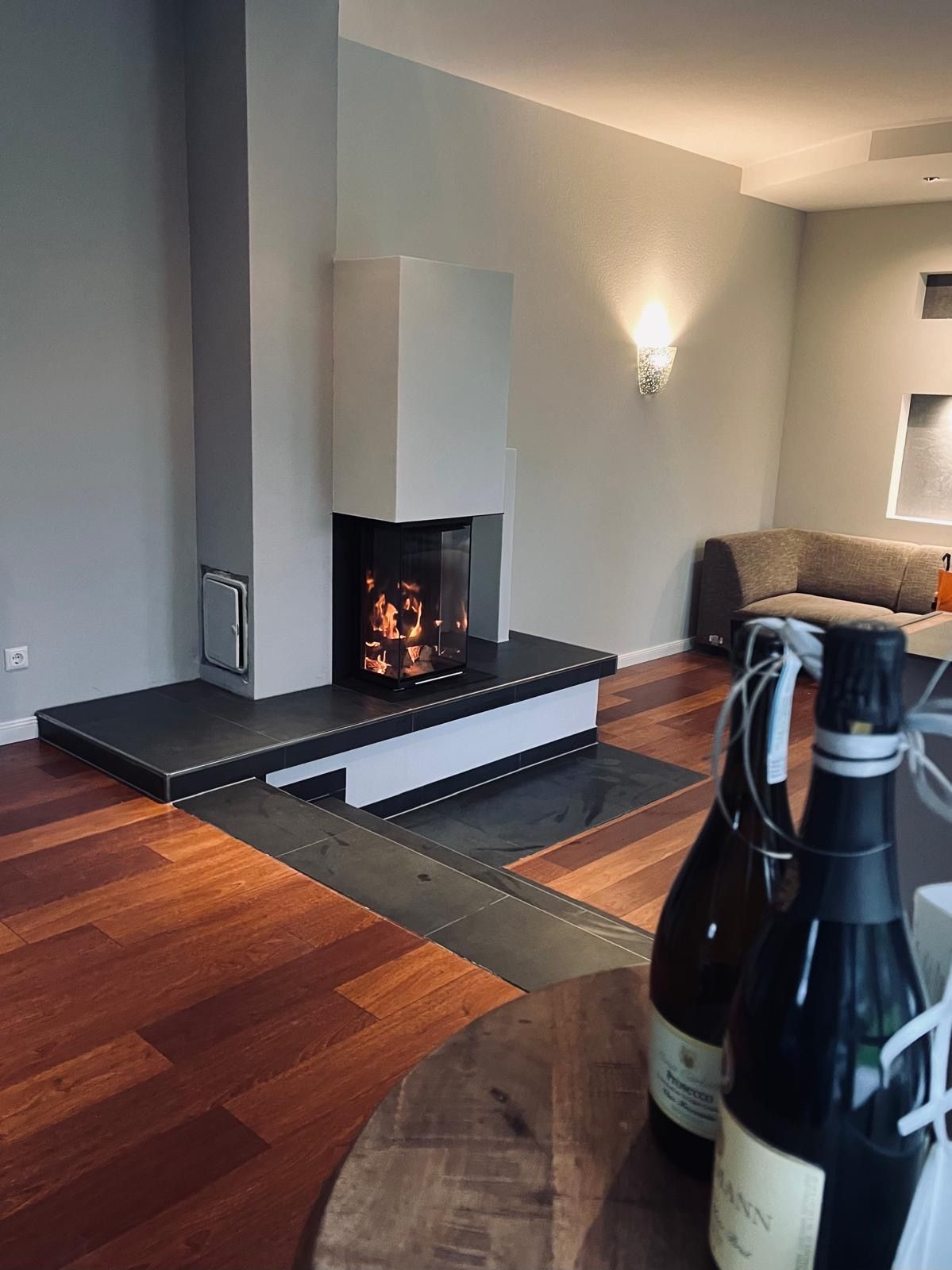 Moderner Kamin mit Flammen in einem grau-weißen Wohnraum; Flaschen im Vordergrund.