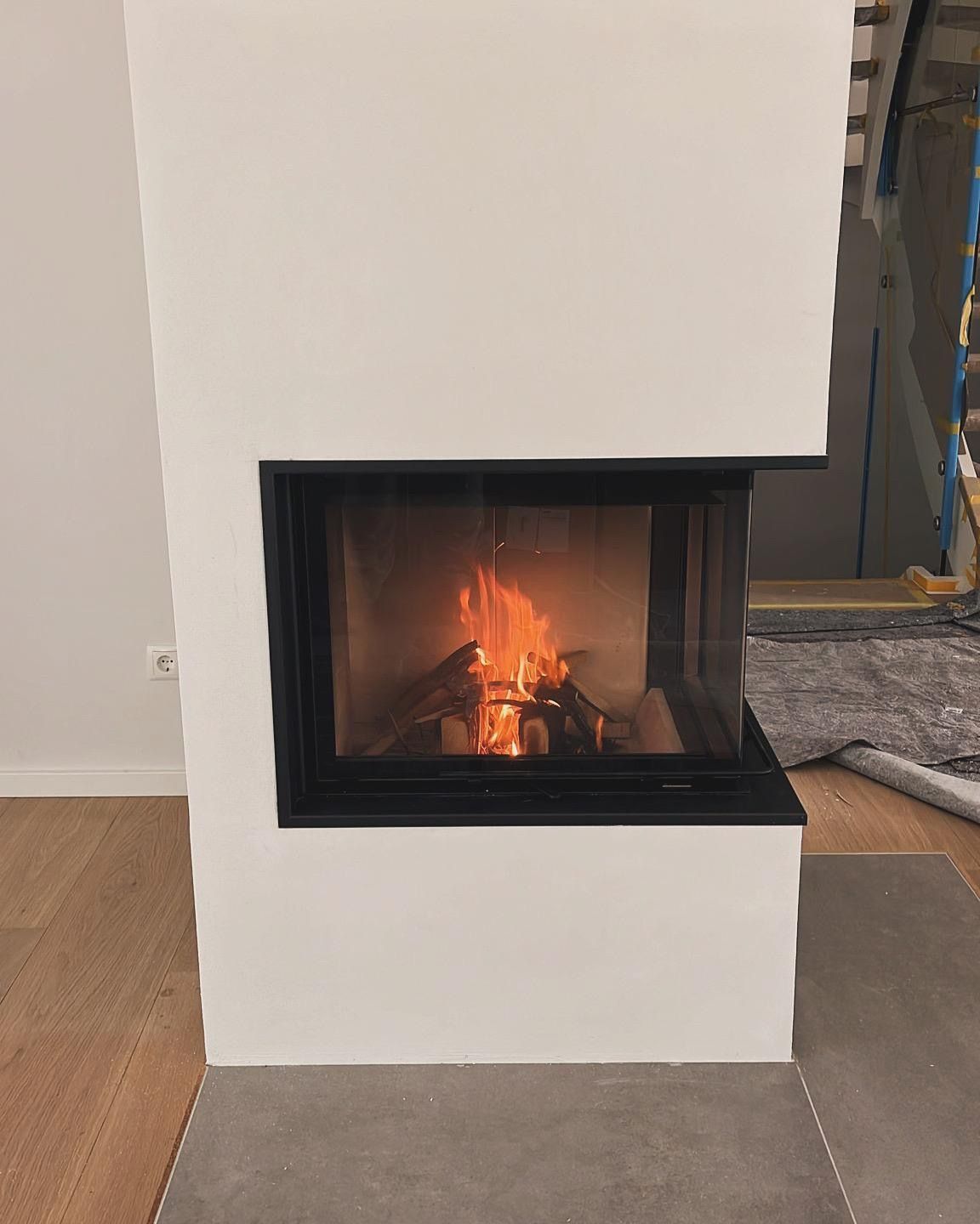 Kamin mit brennendem Holz, schwarzem Rahmen, weißer Umrandung, grauem Boden.