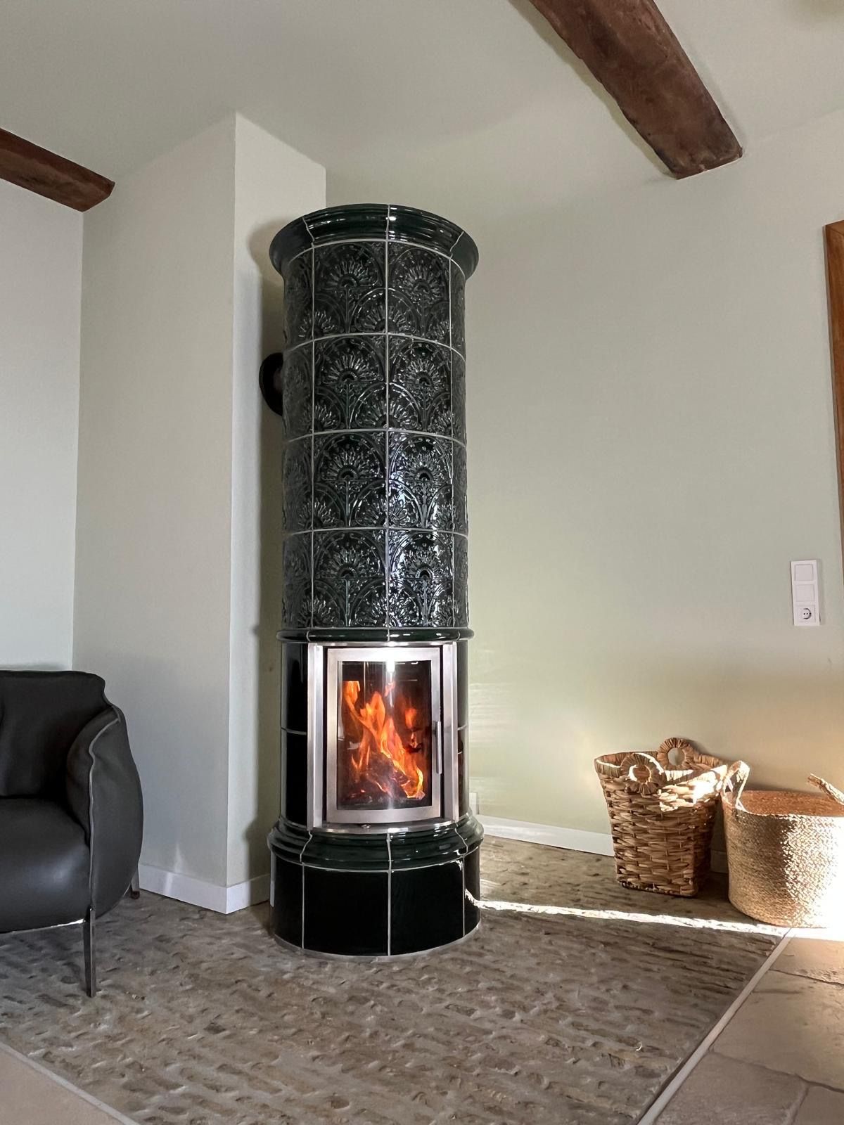 In einem Eckzimmer steht ein grüner Kachelofen mit brennenden Flammen, neben einer schwarzen Ledercouch.