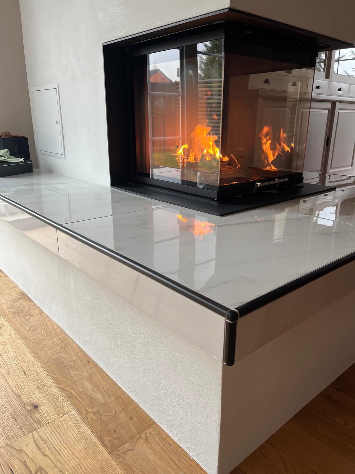Kamin mit Flammen, weißer Marmorherdplatte, schwarzer Umrandung, Eckdesign und hellem Holzboden.