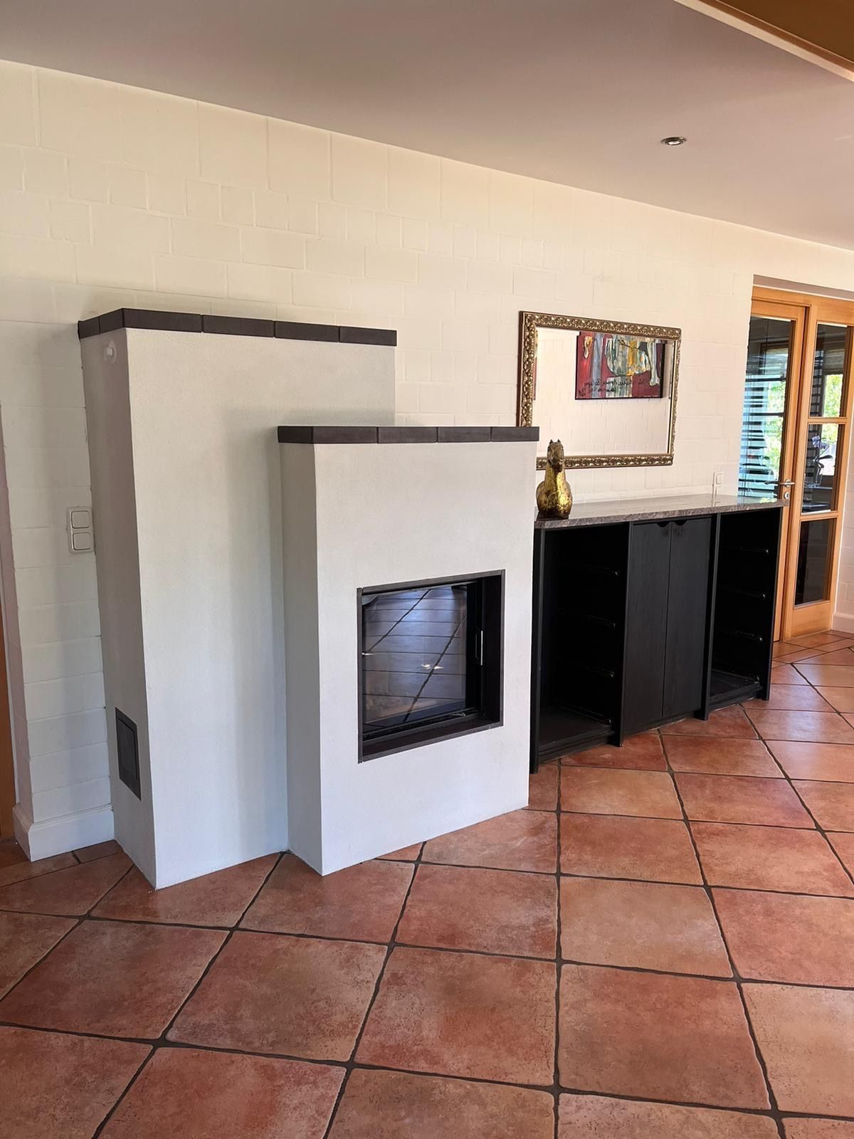 Weißer Kamin mit schwarzem Rahmen, Fliesenboden, schwarzer Schrank, Spiegel und eine Tür mit Jalousien.