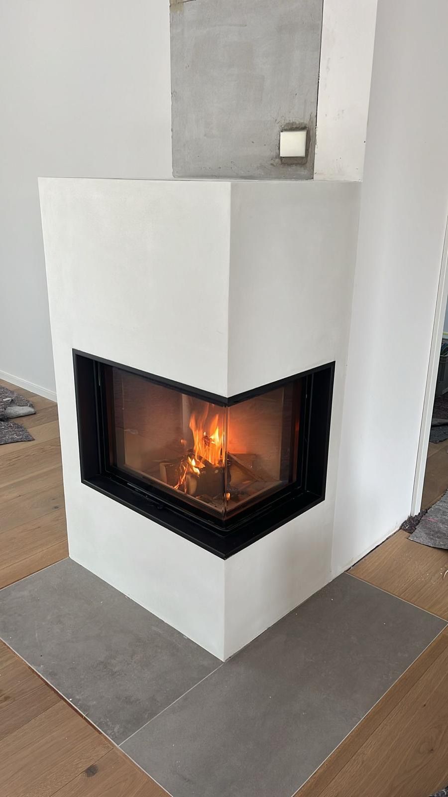 Weißer Eckkamin mit einem hell lodernden, schwarz umrahmten Feuer auf gefliestem Kaminboden.