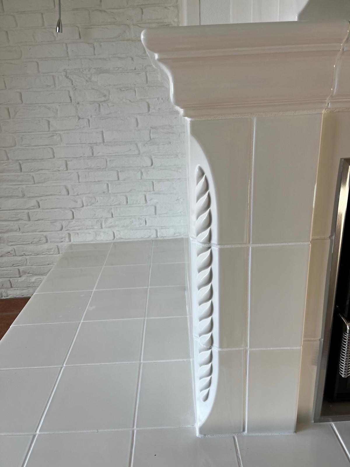 Weiß gefliester Kamin mit dekorativem Lüftungsgitter, neben einer weißen Ziegelwand und gefliester Fläche.