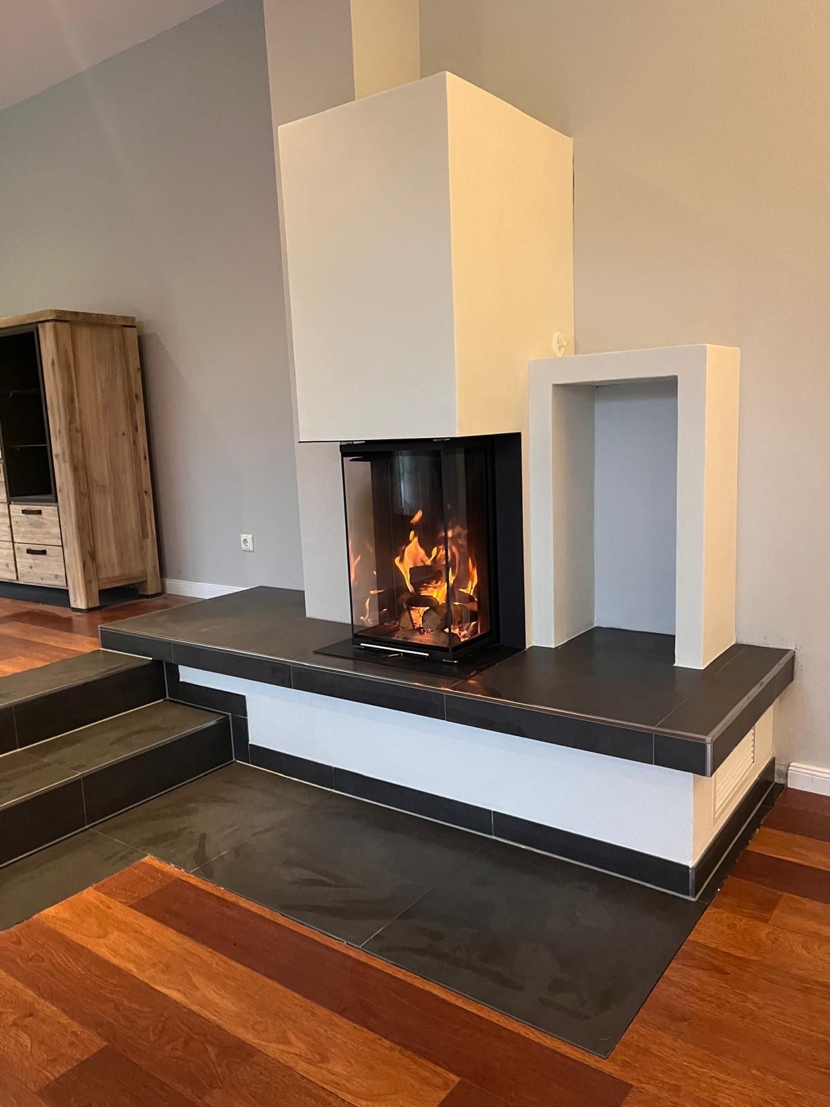 Moderner Kamin mit Glasfront, durch die die Flammen sichtbar sind, auf einem erhöhten Podest mit schwarzen Fliesen. Details in Hellbeige und Weiß.