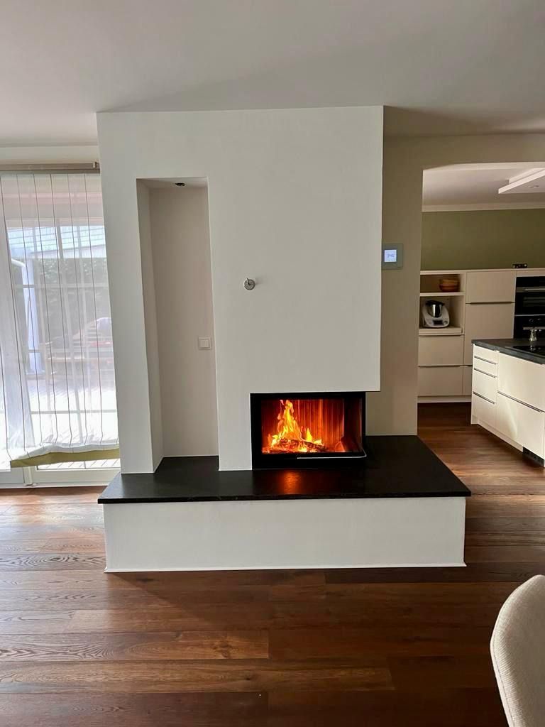 Moderner Kamin mit lodernden Flammen, in eine weiße Wand eingelassen. Dunkle Feuerstelle, Holzboden, Küche im Hintergrund.