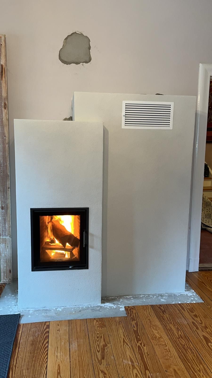 Moderner Kamin mit brennendem Feuer, hellgrauer Fassade und Lüftungsöffnung.