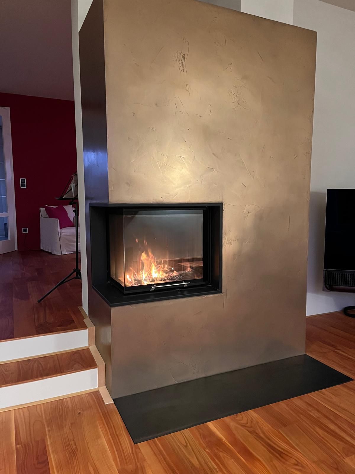 Ein moderner Kamin mit loderndem Feuer in einer bronzefarbenen Wand. Holzboden im Raum.