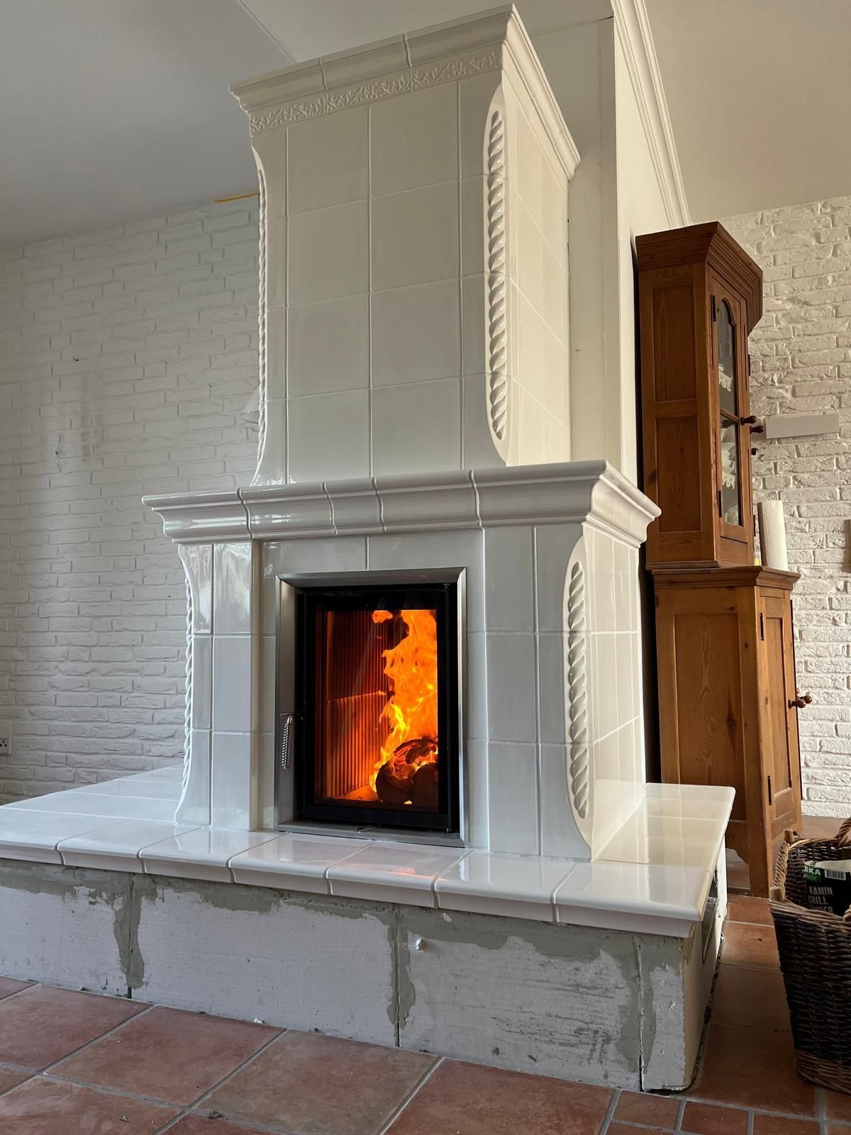 Ein weiß gefliester Kamin mit brennendem Feuer vor einer weißen Backsteinwand und Holzmöbeln.