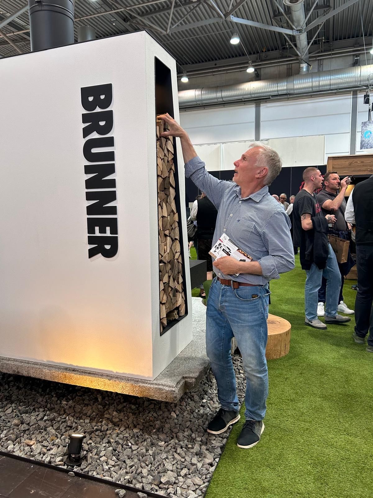 Ein Mann greift in einen weißen Kamin der Marke „BRUNNER“, in dem sich Holzscheite befinden. Andere Personen stehen in der Nähe in einem Veranstaltungsraum.
