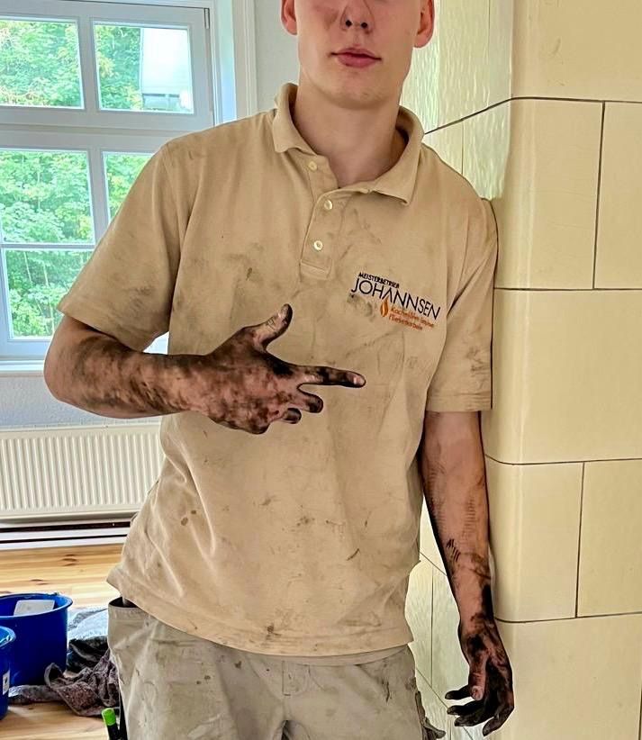 Ein Mann in einem hellbraunen Arbeitshemd, mit Schmutz bedeckt, lehnt an einer weiß gefliesten Wand.