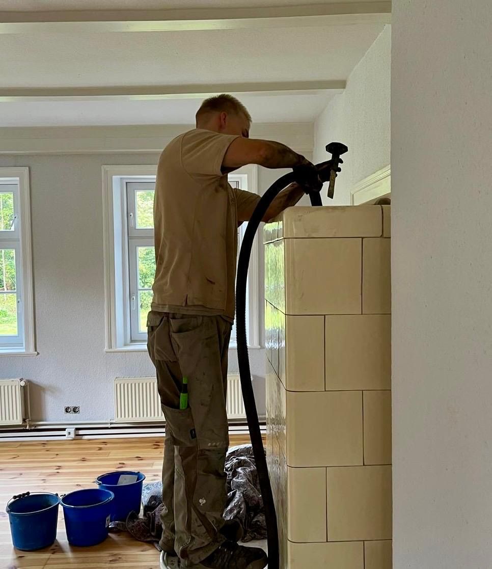 Ein Mann benutzt ein Werkzeug, um in einem Innenraum an einem Backsteingebäude zu arbeiten; blaue Eimer stehen auf dem Boden.