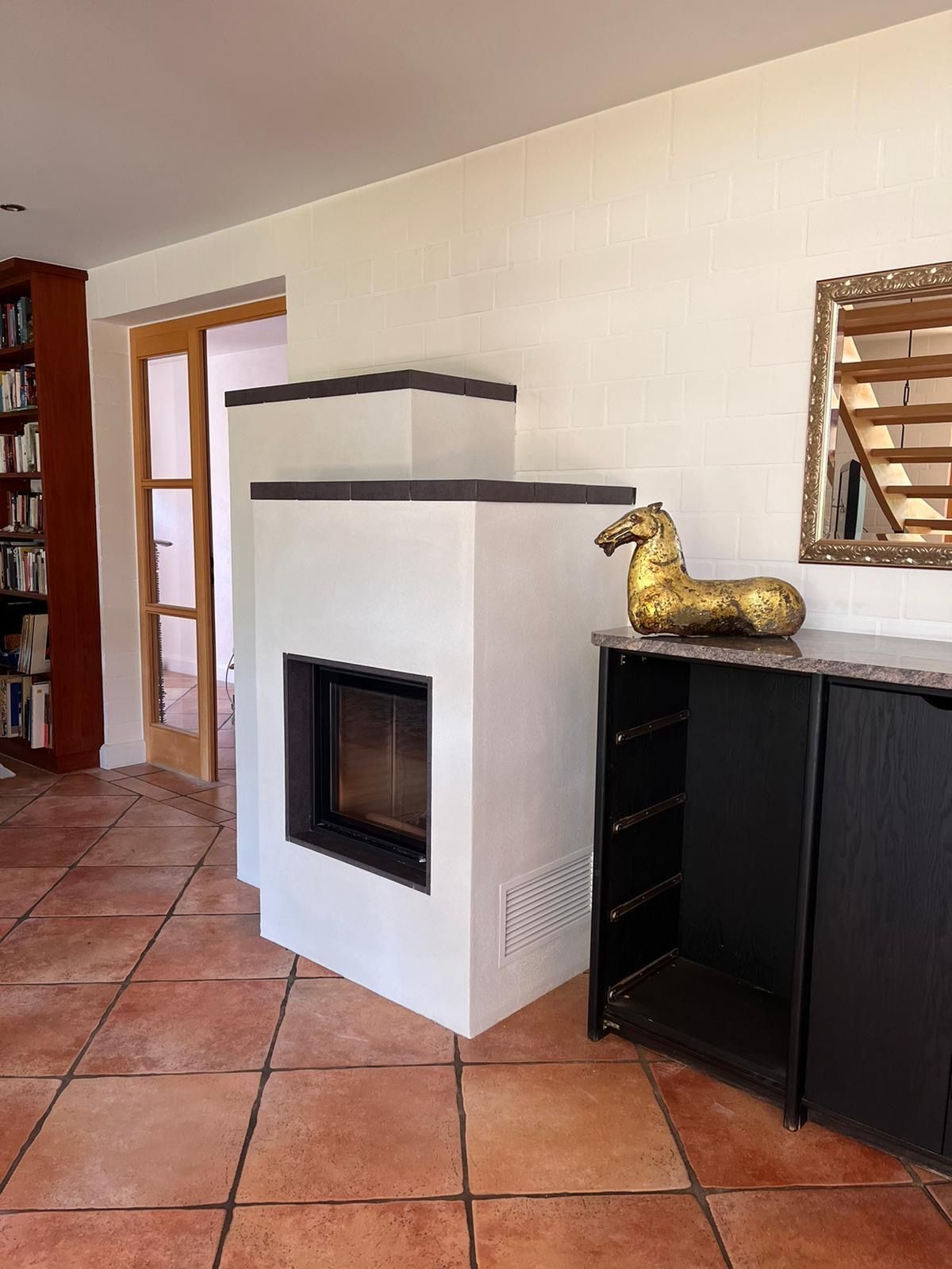 Weißer Kamin mit schwarzem Zierrand, goldene Pferdestatue auf schwarzem Schrank, Terrakottafliesenboden.