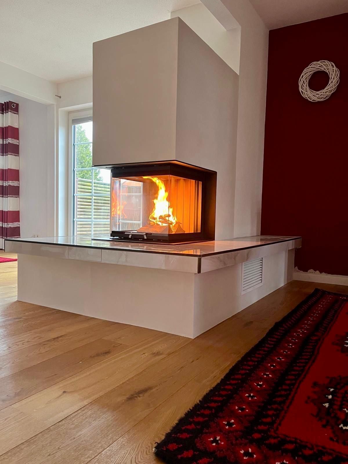 Ein moderner Kamin mit brennendem Feuer, weißer Konstruktion und Glasfront, in einem Raum mit Holzboden und roter Wand.