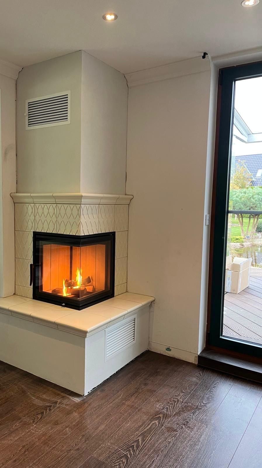 Kamin in der Zimmerecke mit brennenden Flammen. Weiße Fliesenfassade, brauner Boden, Glastür zur Terrasse.