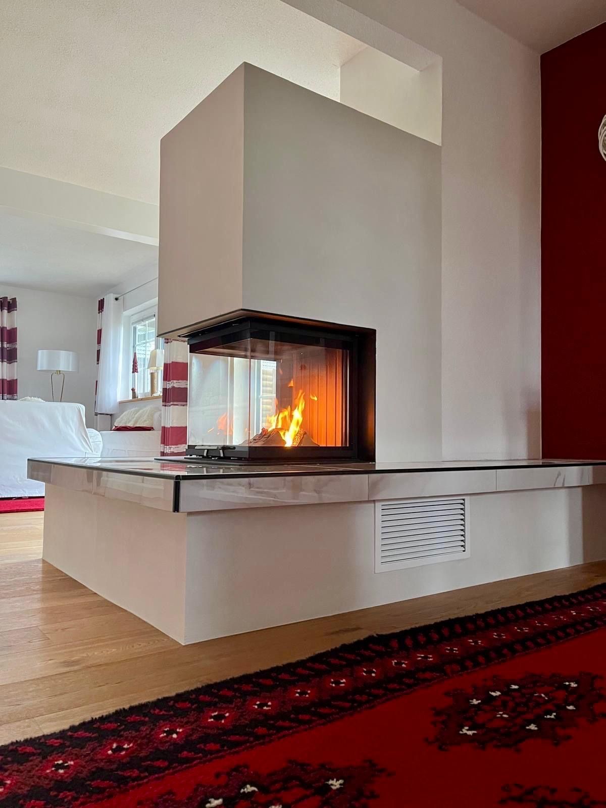 Moderner Kamin mit brennendem Feuer, weißes Gestell, Glasfront. Roter Teppich und rote Wand.