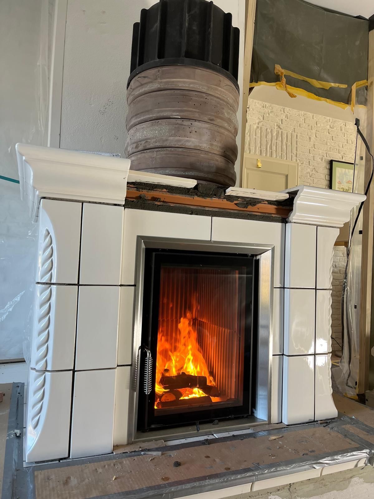 Kamin mit weiß gefliester Umrandung, brennende Flammen hinter einer Glastür sichtbar, schwarzer Schornstein.