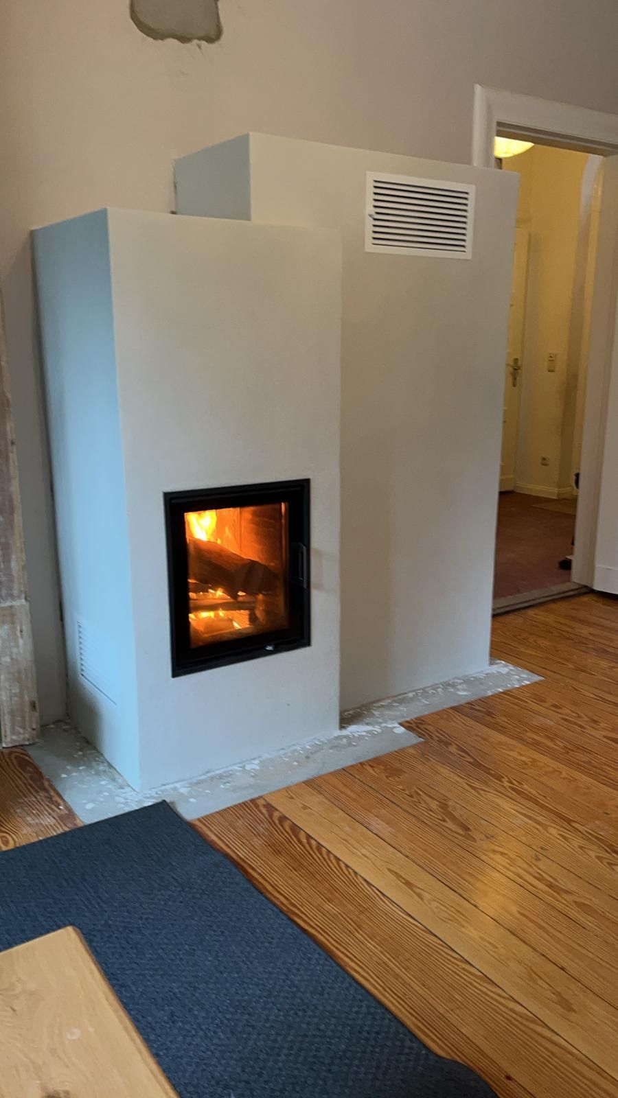 Ein hellgrauer, moderner Kamin mit brennendem Feuer. Davor befindet sich ein Holzboden und ein blauer Teppich.