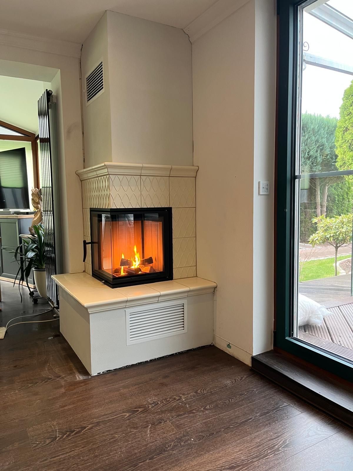 Weißer Kamin mit Feuer, neben einer Glastür mit Blick auf einen Garten.