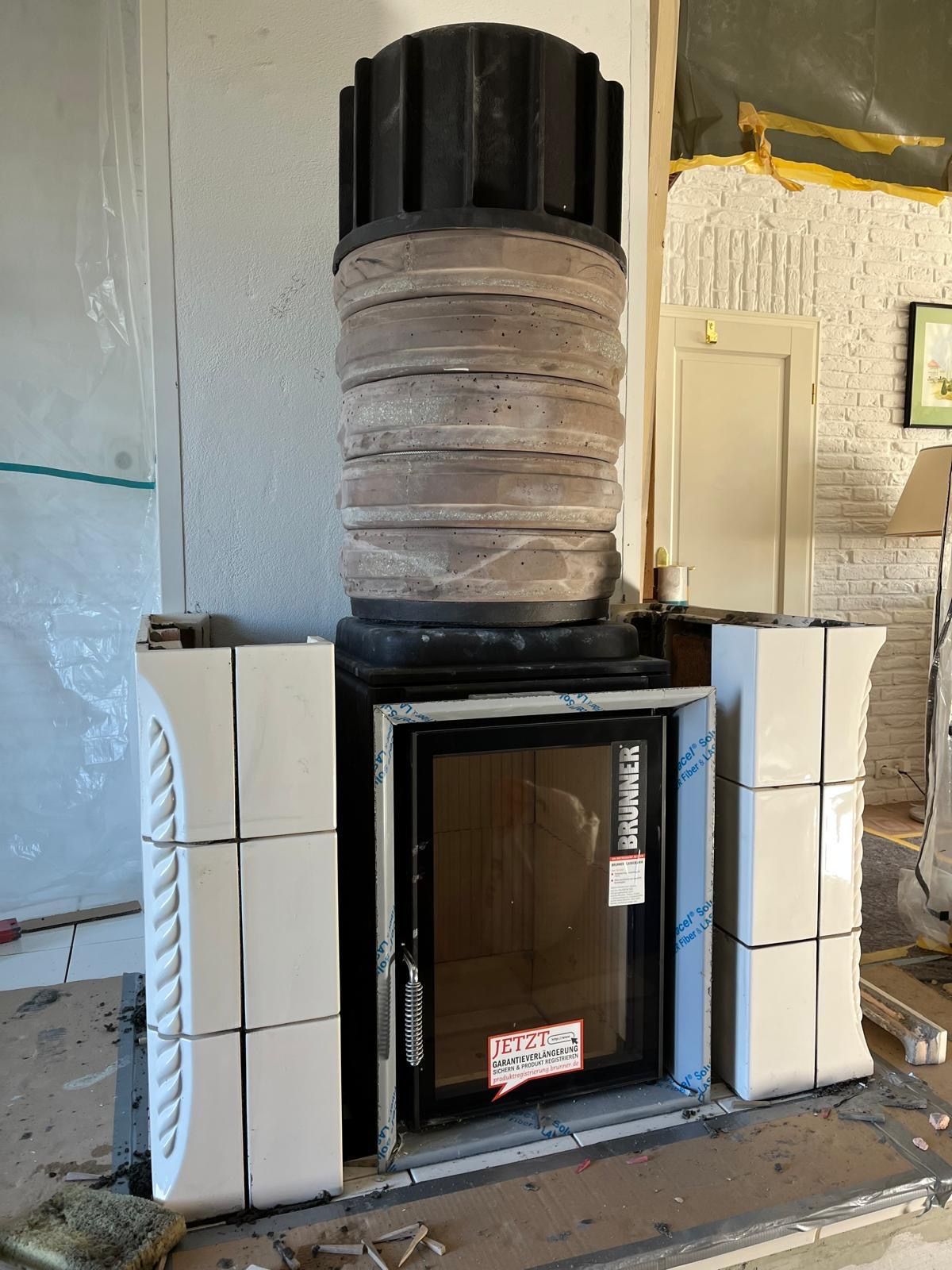 Kamin im Bau, schwarze Tür, weiße Fliesenumrandung, grauer Schornstein, an einer Wand.