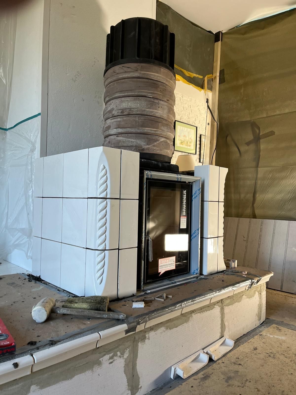 Weißer Kachelofen im Bau, mit schwarzem Schornstein und Sichtfenster.