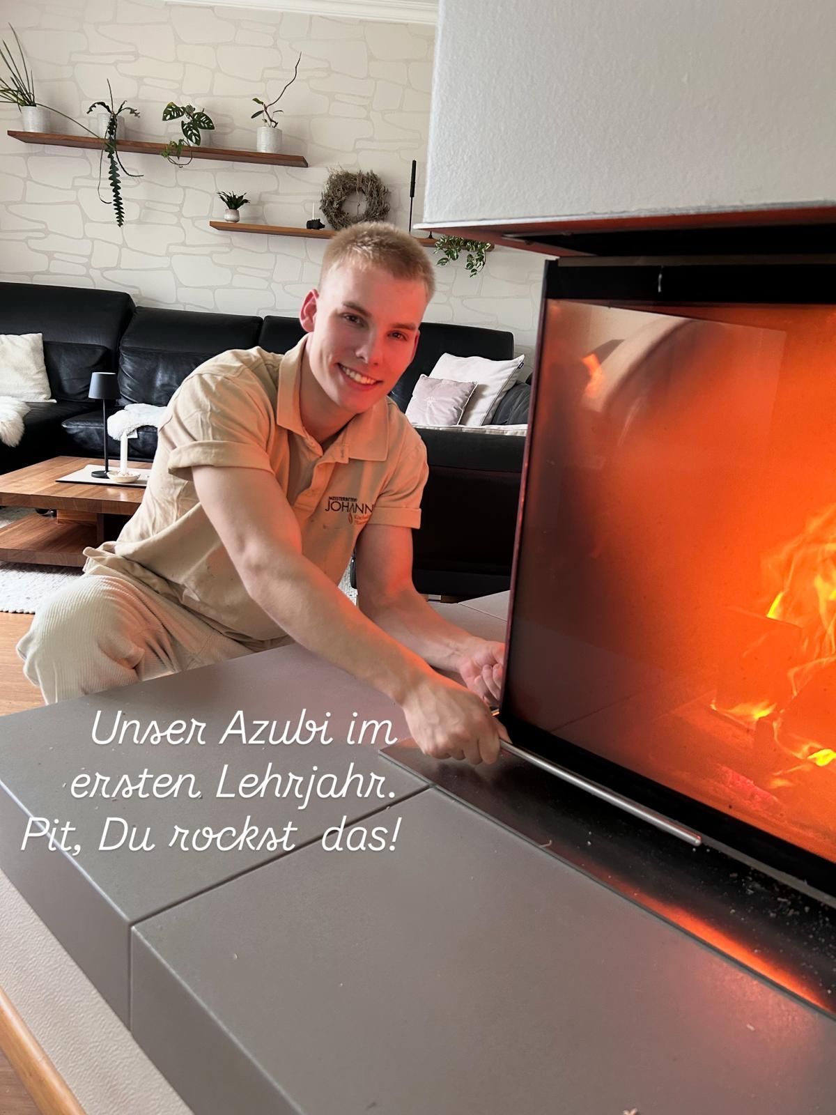 Lehrling arbeitet am Kamin. Mann lächelt, Kaminflammen sind sichtbar. Zimmer mit Regalen und Sofa.