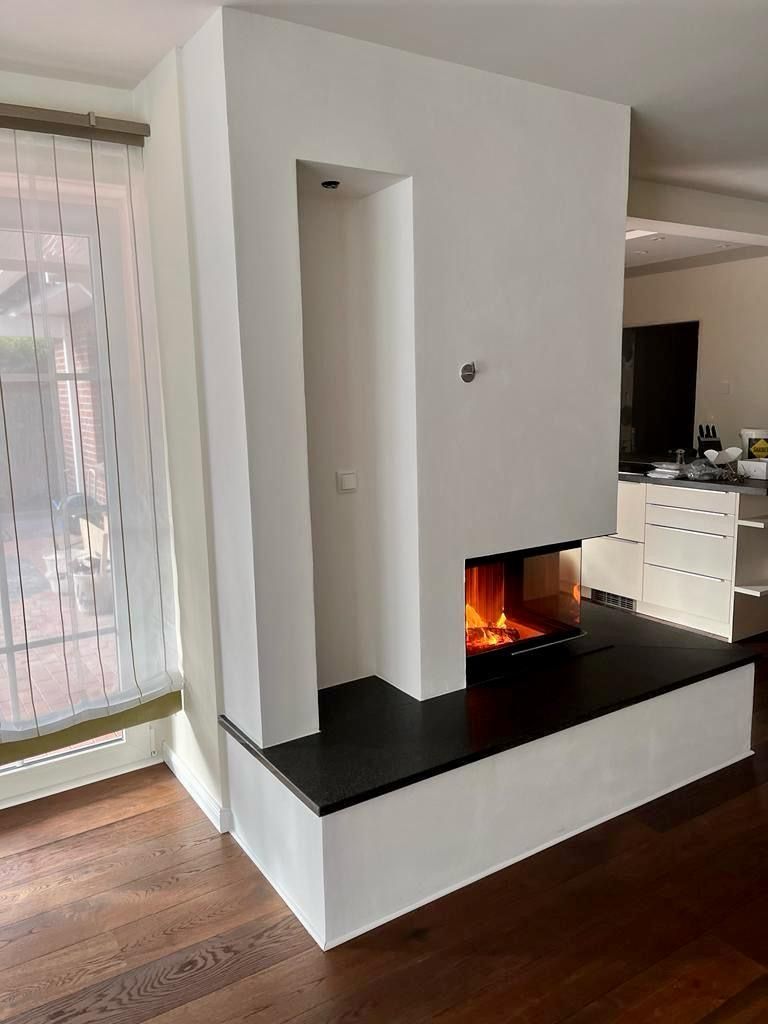 Moderner weißer Kamin mit schwarzer Abdeckung. Innenraum mit Holzboden und Fenster. Feuer brennt im Inneren.