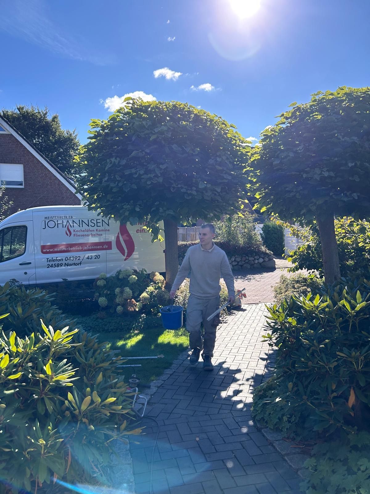 Ein Mann mit Werkzeugen geht auf einem Backsteinweg auf die Kamera zu; im sonnigen Hintergrund sind ein Lieferwagen und Bäume zu sehen.