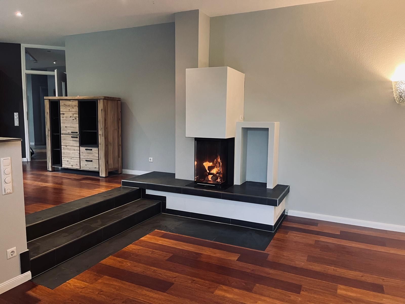 Wohnzimmer mit Kamin, Treppe und Holzboden. Feuer im Kaminsims, neutrale Wände, dunkler Holzschrank.