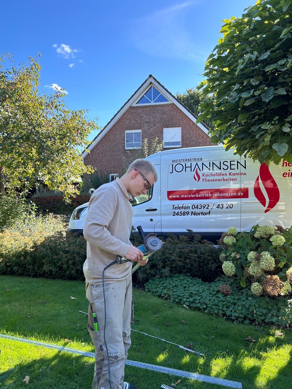 Ein Mann benutzt ein Werkzeug vor einem weißen Lieferwagen mit Firmenlogo. Im Hintergrund sind ein Haus und Bäume zu sehen.