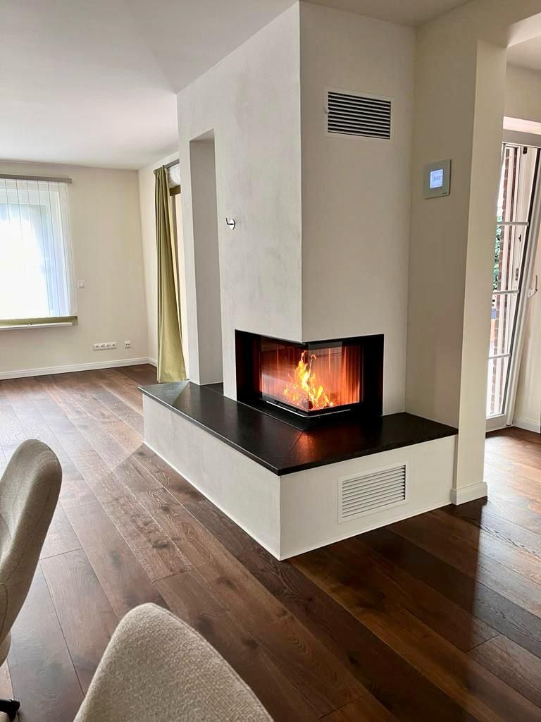 Eckkamin mit Feuerstelle, eingefasst in eine weiße Konstruktion, auf einem dunklen Steinsockel. Parkettboden.