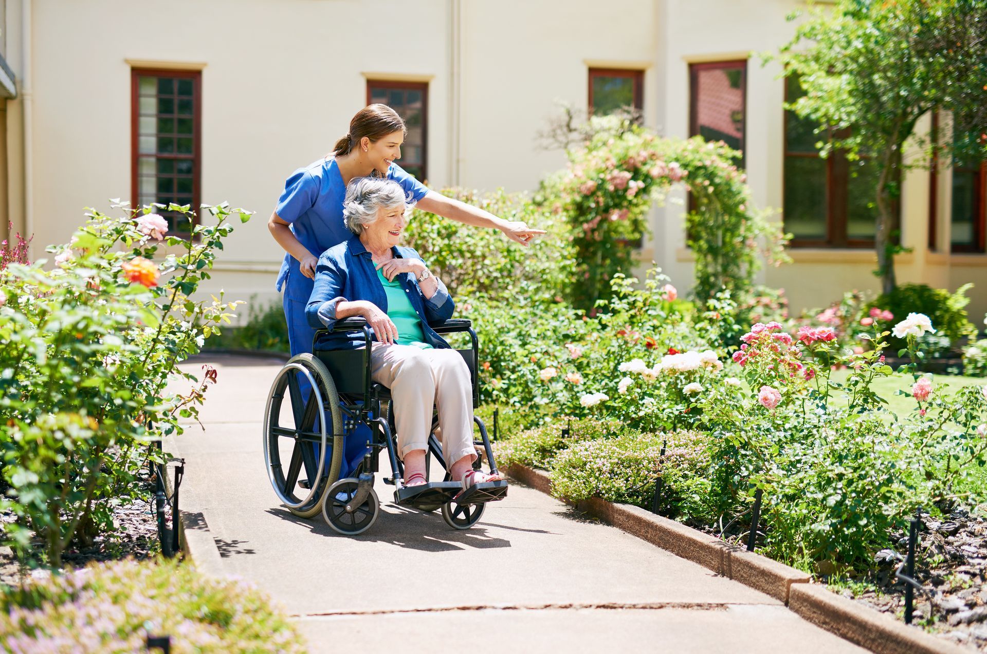 Eine Pflegekraft in blauer OP-Kleidung schiebt eine Person im Rollstuhl einen Gartenweg entlang und zeigt auf die Blumen.