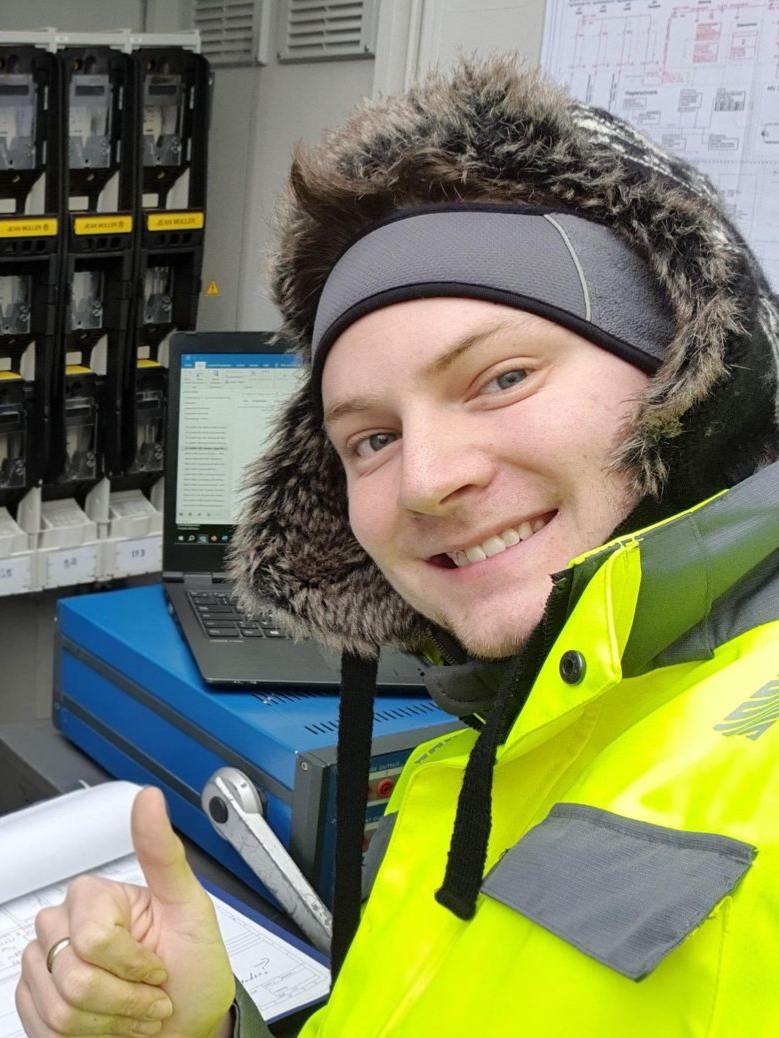 Ein Mann in gelber Jacke und Pelzmütze zeigt neben Ausrüstung und einem Laptop den Daumen nach oben.