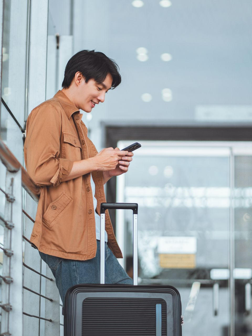 Un homme à l'aéroport, appuyé contre un mur, regarde son téléphone, ses bagages à proximité.