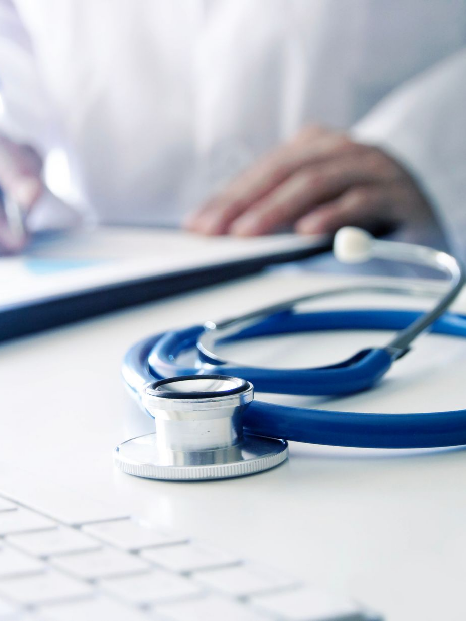 Stéthoscope bleu sur un bureau blanc ; un médecin en arrière-plan écrit sur un bloc-notes.
