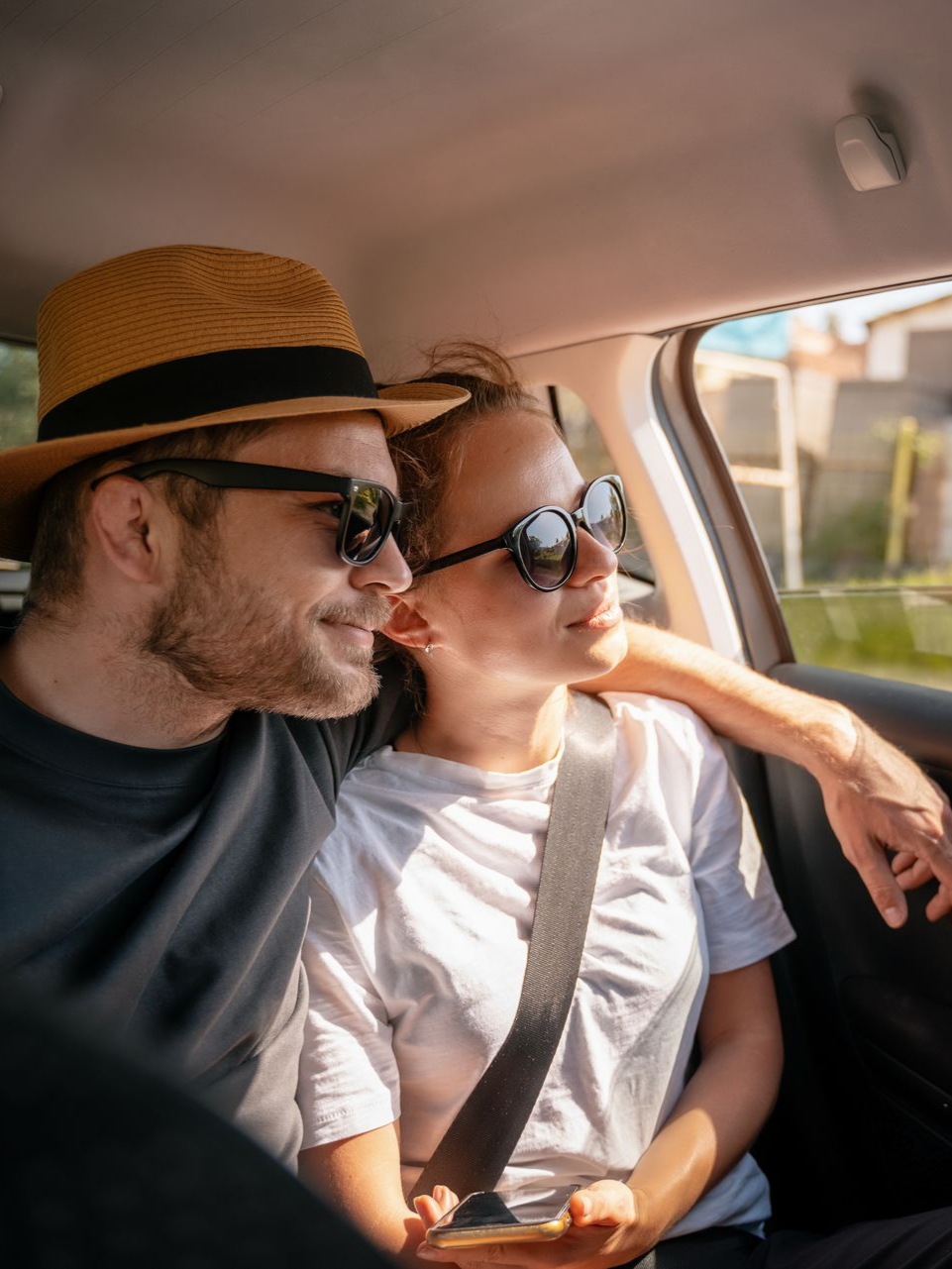Un couple en voiture, regardant par la fenêtre ; l'homme porte un chapeau et des lunettes de soleil, la femme porte des lunettes de soleil.