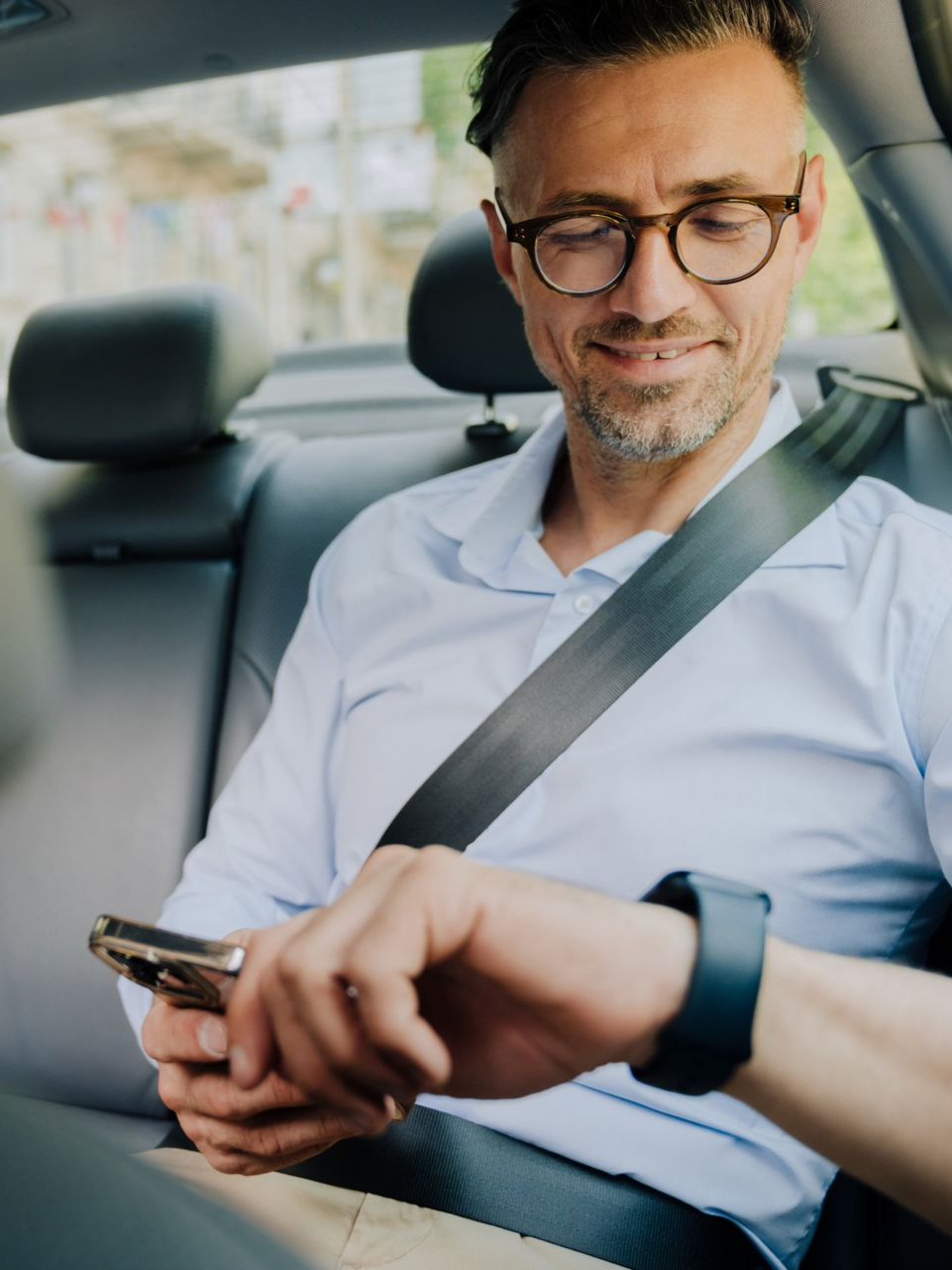 Un homme dans une voiture, portant des lunettes et une chemise bleu clair, regarde une montre connectée tout en tenant un téléphone.