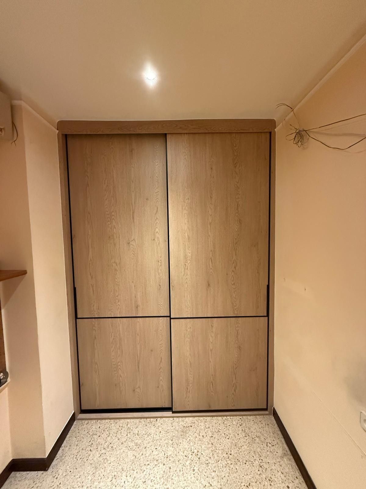 Habitación pequeña de color beige con puertas de armario de madera cerradas y suelo moteado.