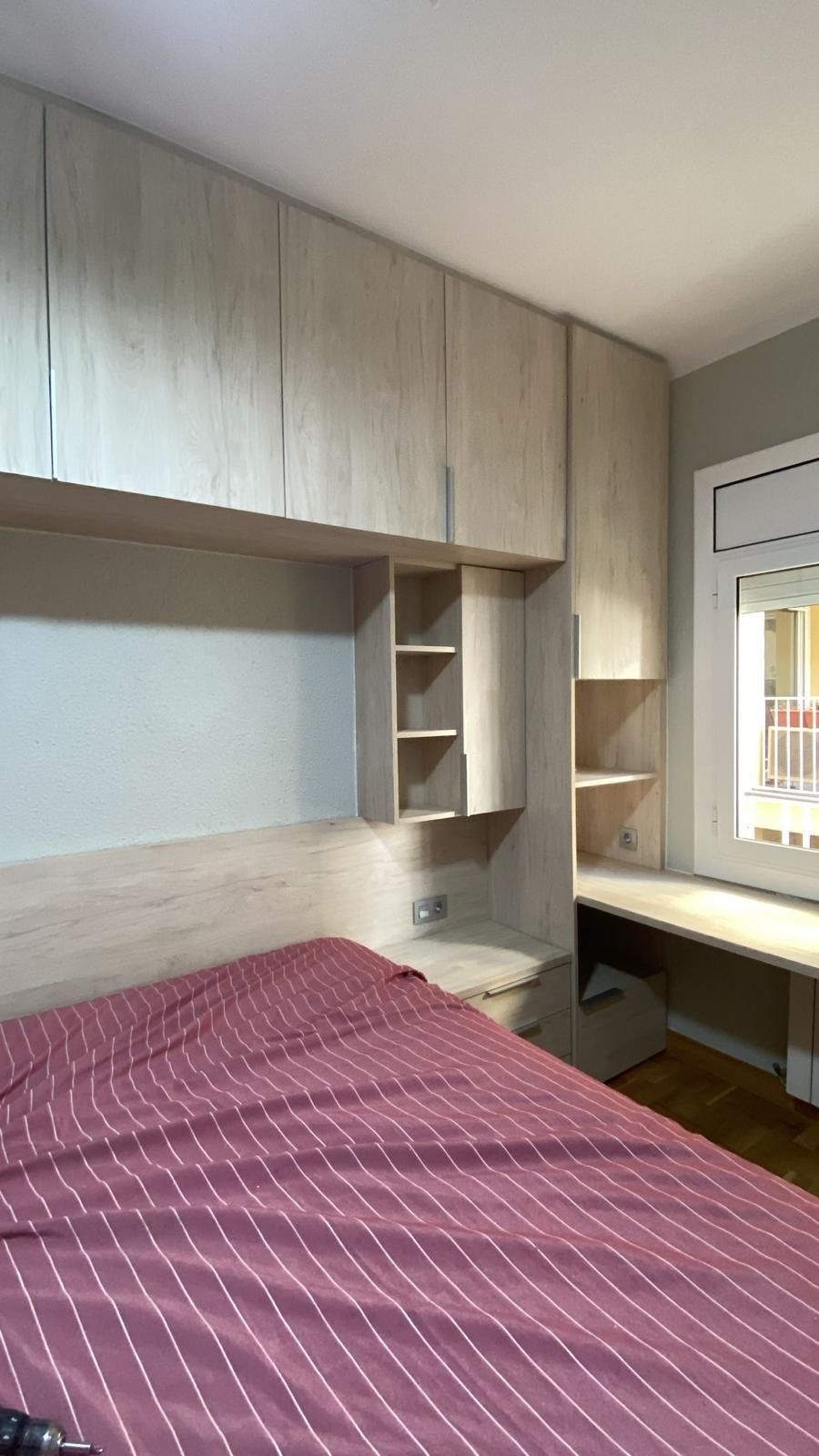 Habitación pequeña con cama, armarios empotrados de madera y un escritorio junto a la ventana.
