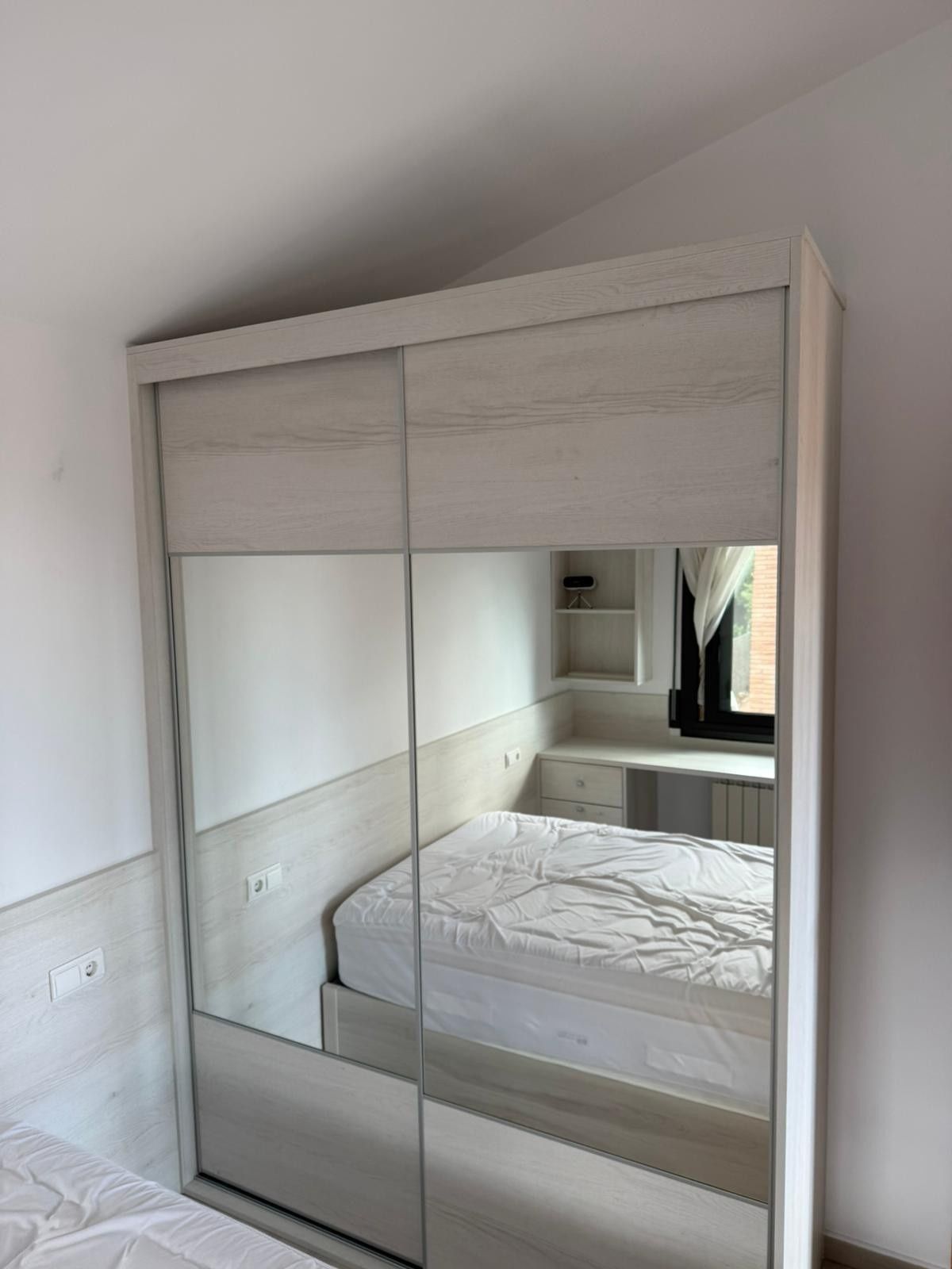 Armario corredero con espejo en un dormitorio luminoso con una cama reflejada en las puertas.