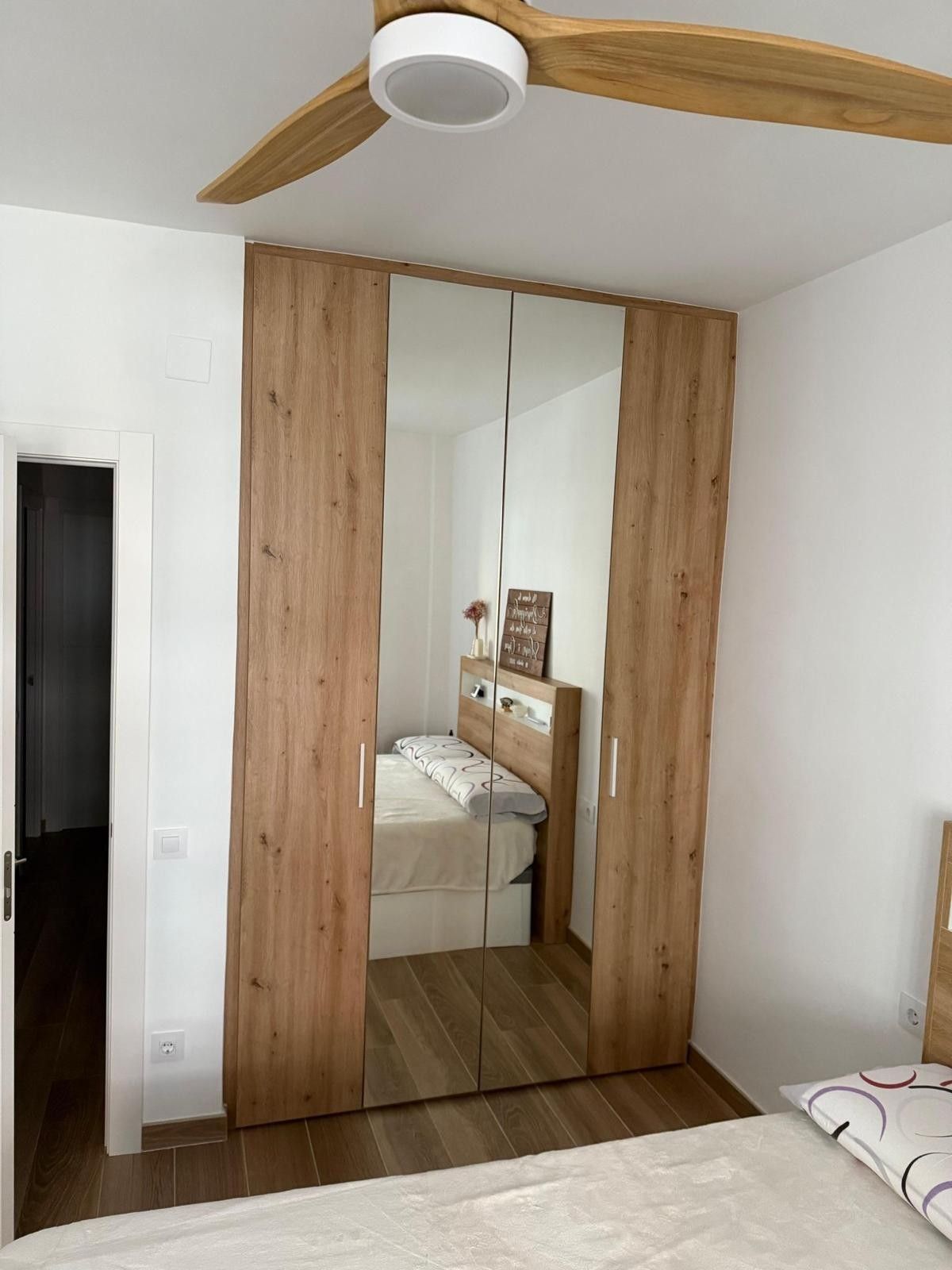 Habitación pequeña con armario de madera, puertas con espejo y ventilador de techo sobre la cama.