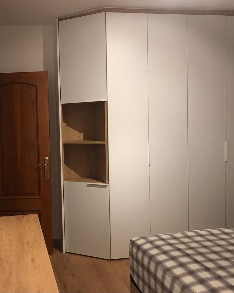 Armario esquinero blanco en un dormitorio con un estante lateral parcialmente abierto y una cama a cuadros en primer plano.