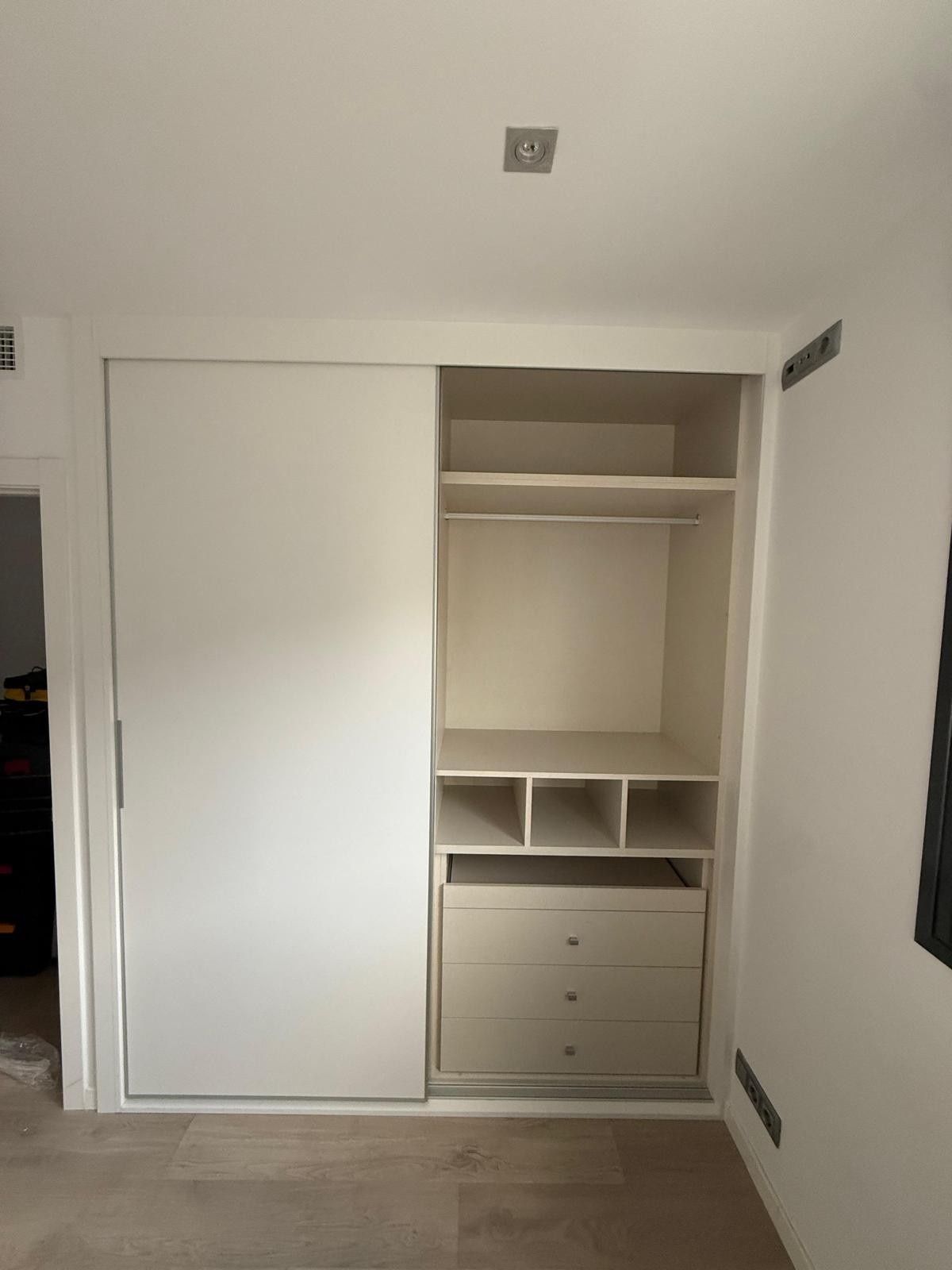 Armario abierto con puerta corredera blanca, estantes y cajones en una habitación blanca.