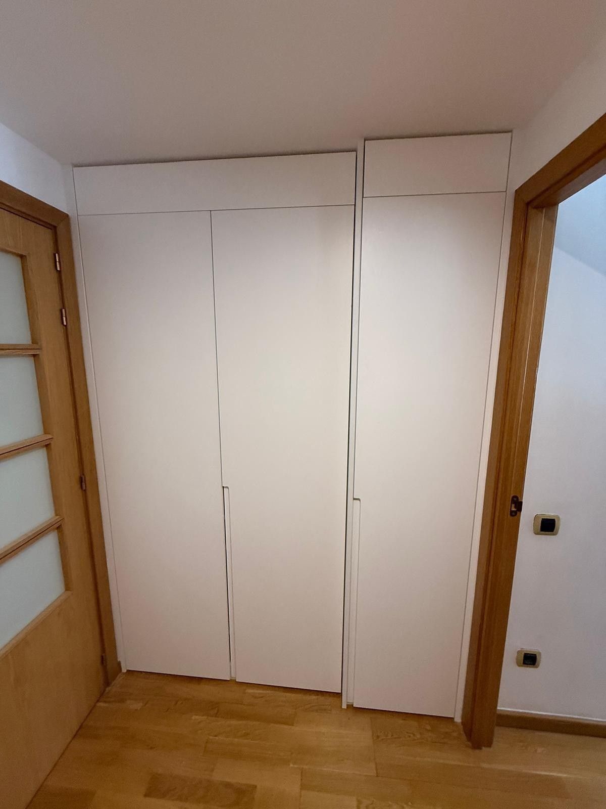 Puertas blancas de armario empotrado entre dos marcos de puertas de madera en una habitación pequeña con suelo de madera.