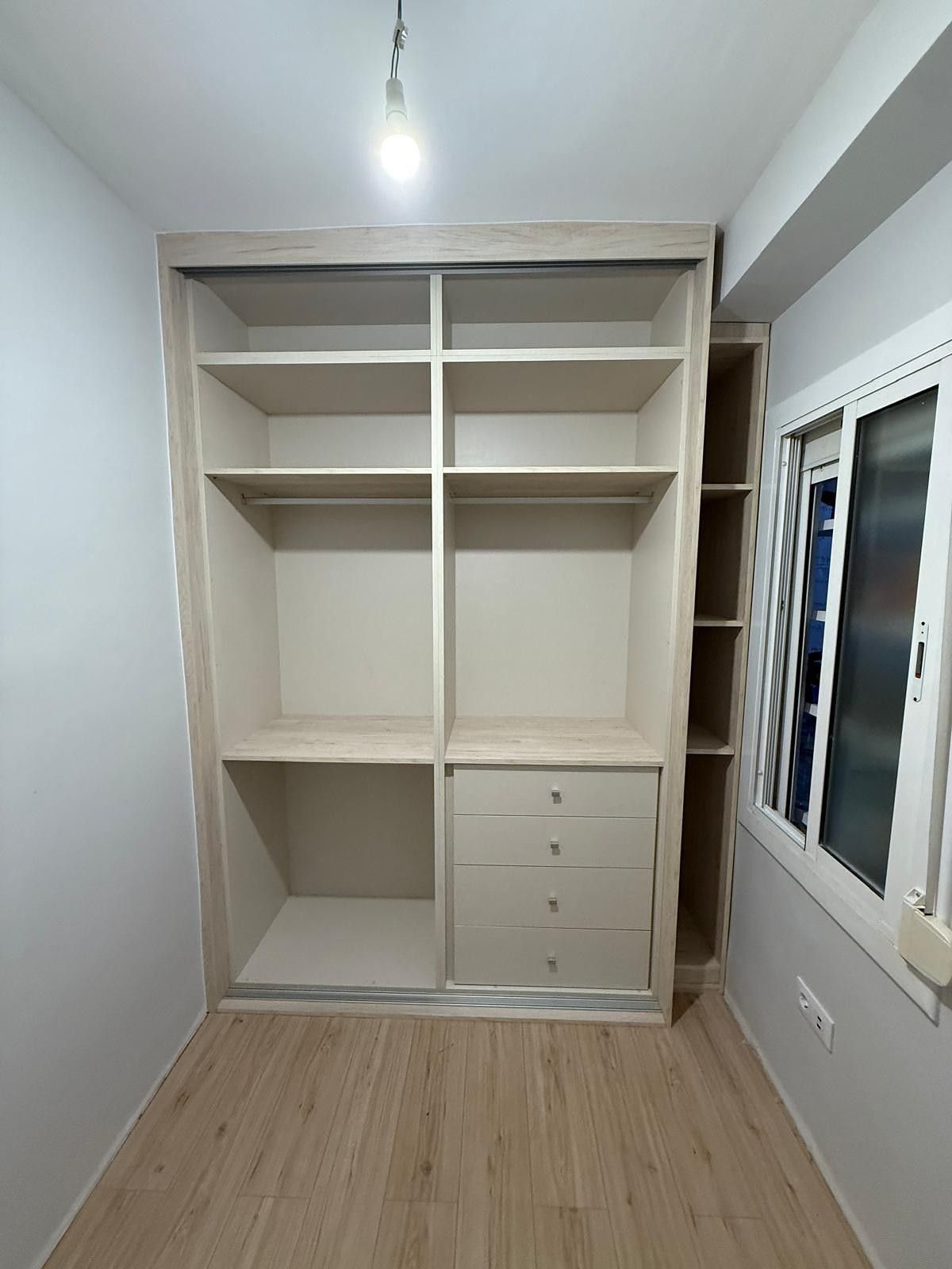 Armario empotrado vacío en una habitación estrecha con estantes blancos, cajones y una ventana a la derecha.