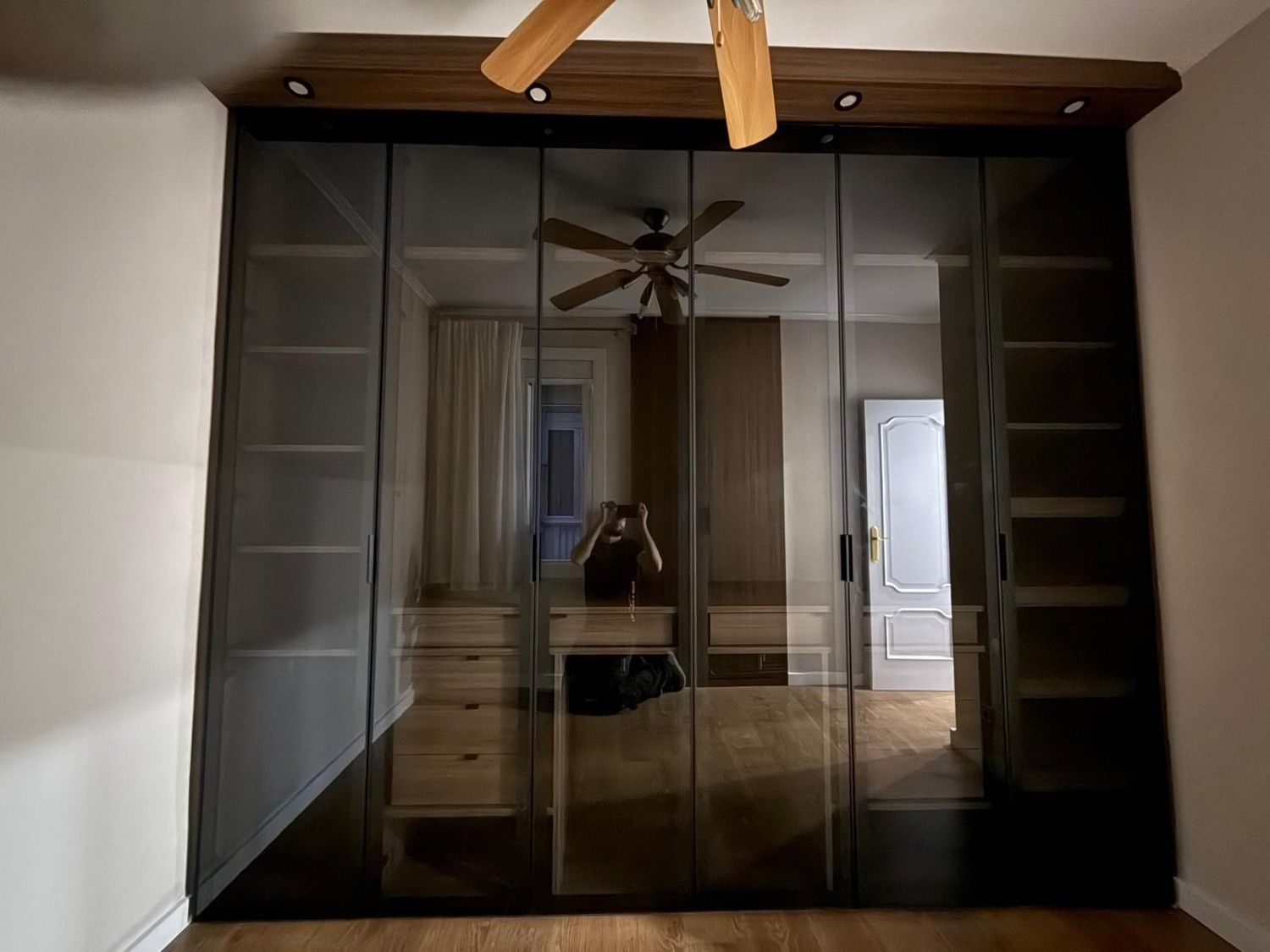 Puertas de armario con espejos y marcos negros en un dormitorio, con el reflejo de un ventilador de techo.